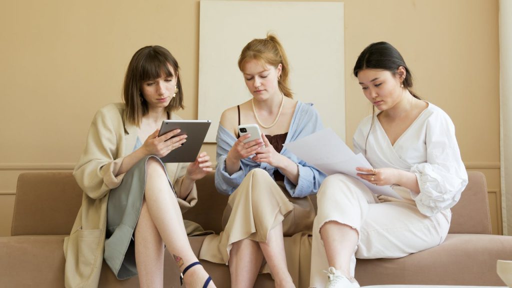 Three women using technology and documents collaborate indoors, leveraging diverse devices for work.
